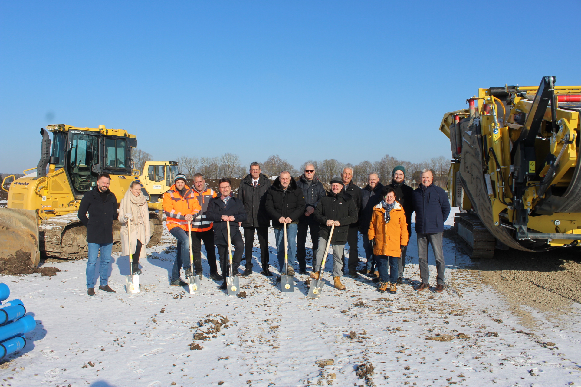 Personen stehen mit Spaten in der Hand auf einer Baustelle mit Baumaschinen im Hintergrund